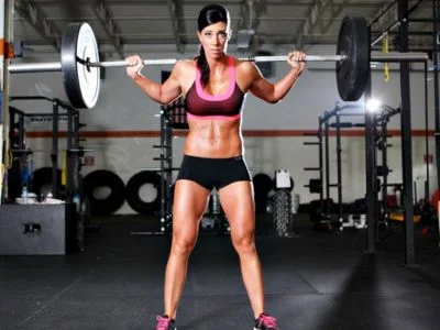 una mujer mirando la camara con una barra en su hombros por hacer sentadillas